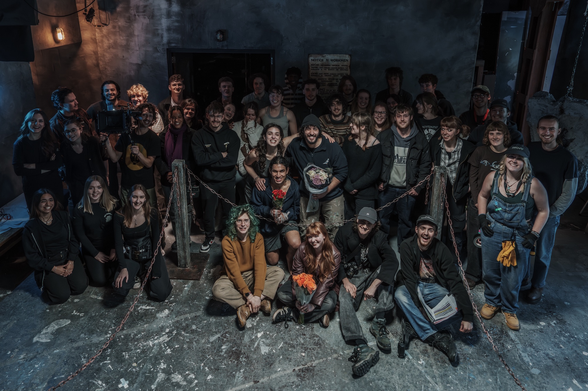 A film crew of approximately 40 people poses together in a dimly lit room. They are arranged in two rows in front of a dark wall and a door, with some individuals standing and others seated on the ground.