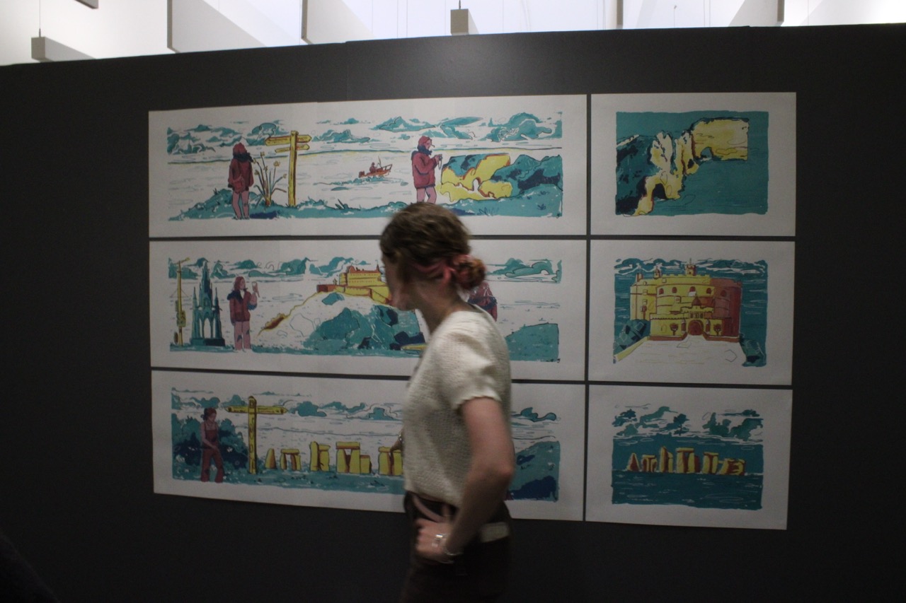 A woman walks past a wall display of eight framed landscape sketches, arranged in a grid on a dark background.