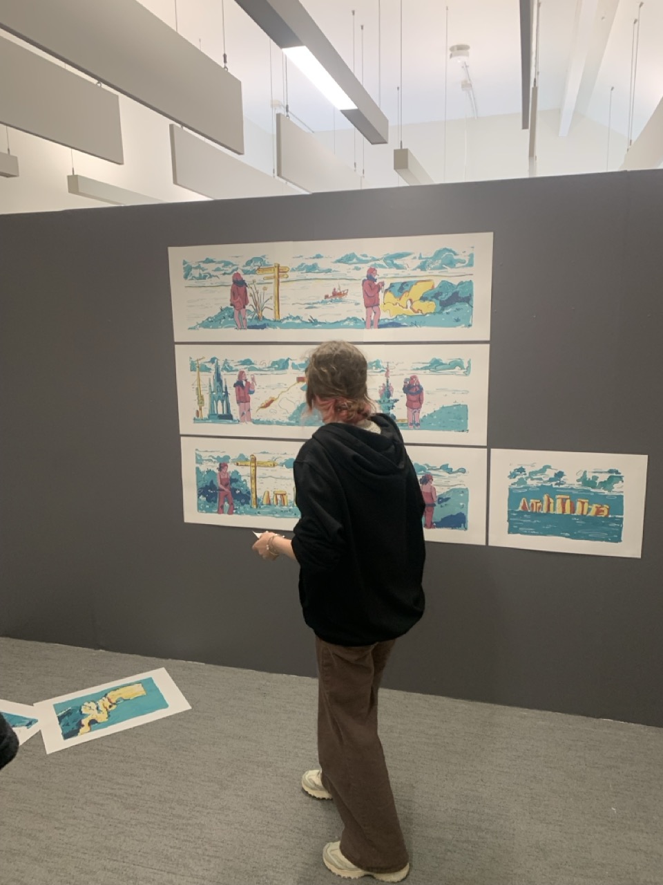 A person stands in a gallery beside a dark display wall showing several colourful rectangular illustrations mounted in rows, with additional prints on the floor and overhead lights above.