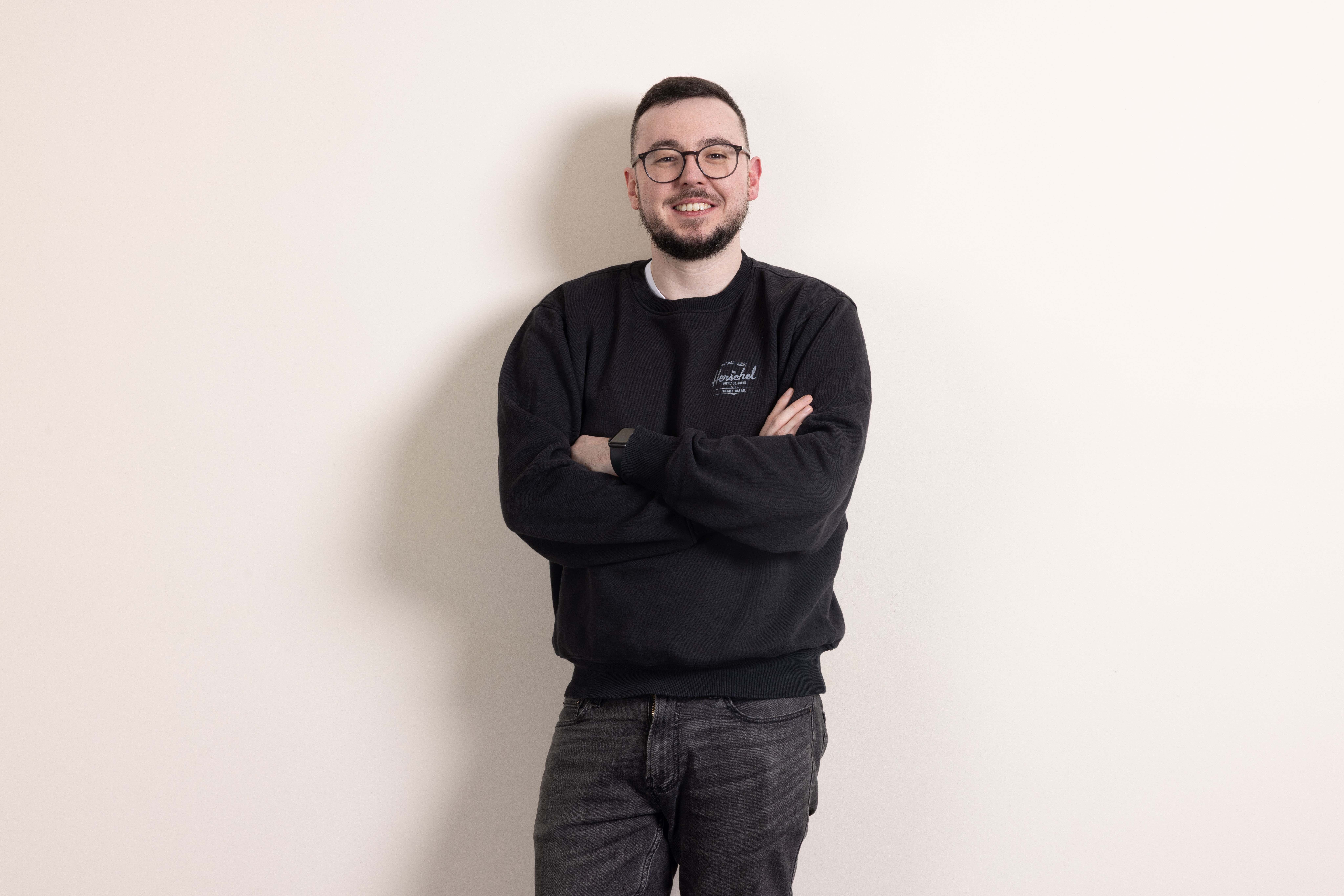 Smiling man with glasses wearing black jumper and grey jeans, arms crossed, standing against white wall.