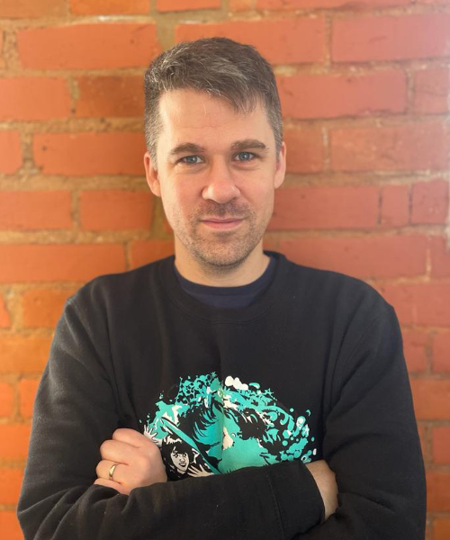 Man with short brown hair wearing black jumper with teal graphic design, arms crossed, standing against red brick wall.