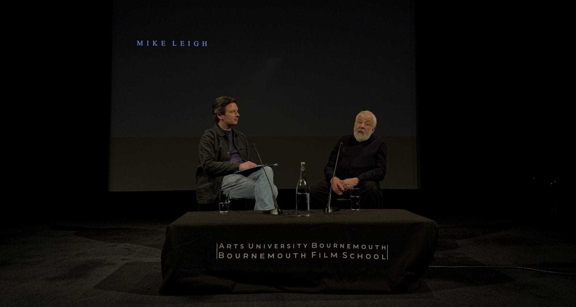Two people sit on a stage before a table and speak. There are microphones standing on the table.
