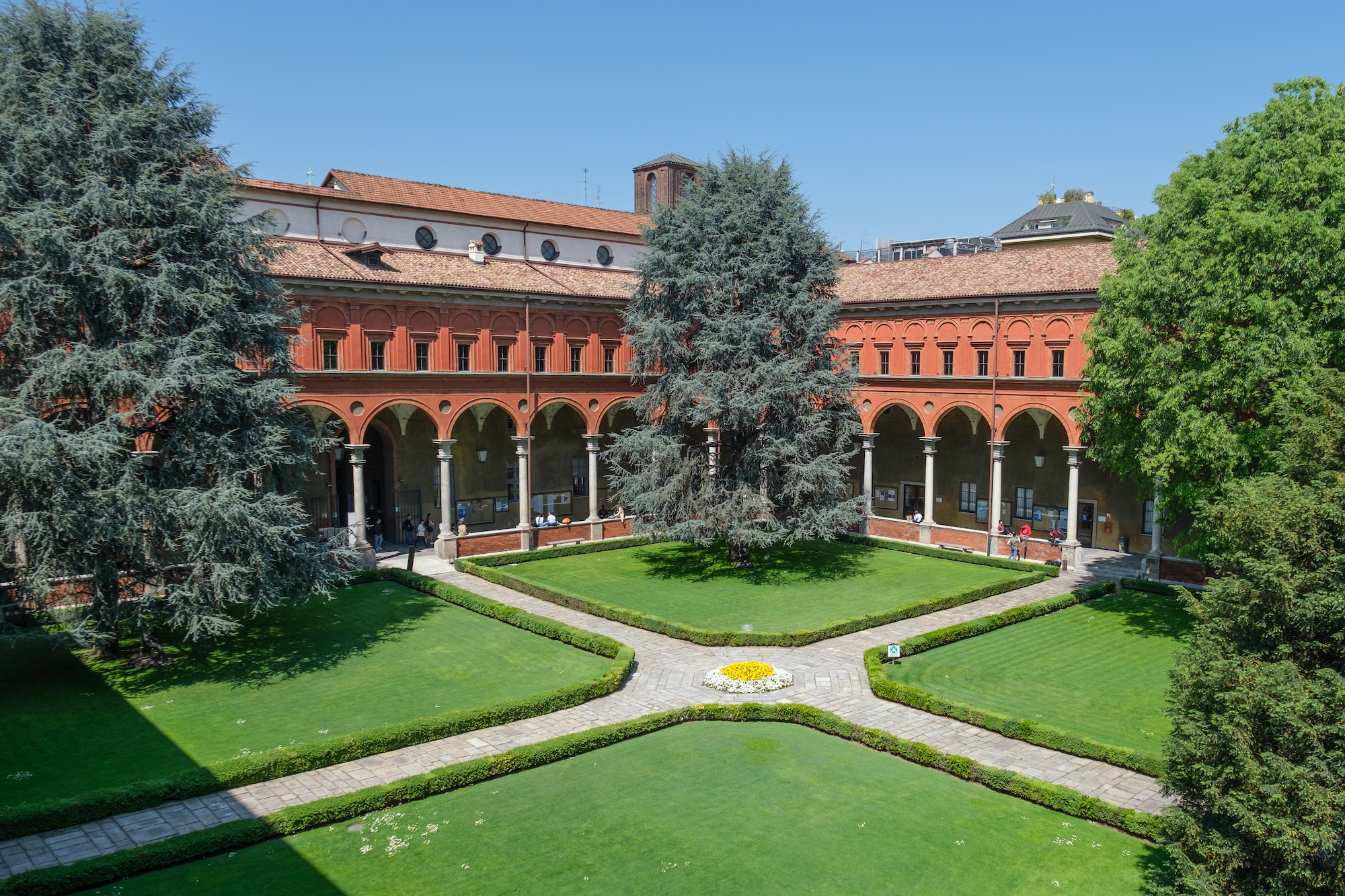 A university courtyard with a cross path, green lawns and trees, and red brick cloisters.
