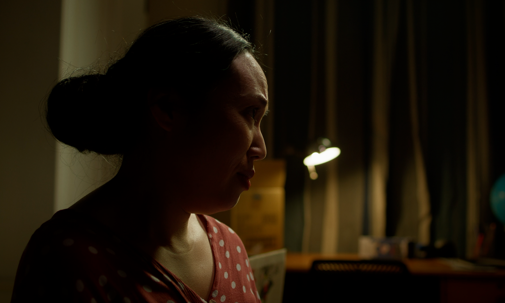 Person in profile with hair in bun wearing polka dot top, lit by warm light against blurred background of wooden shelving.