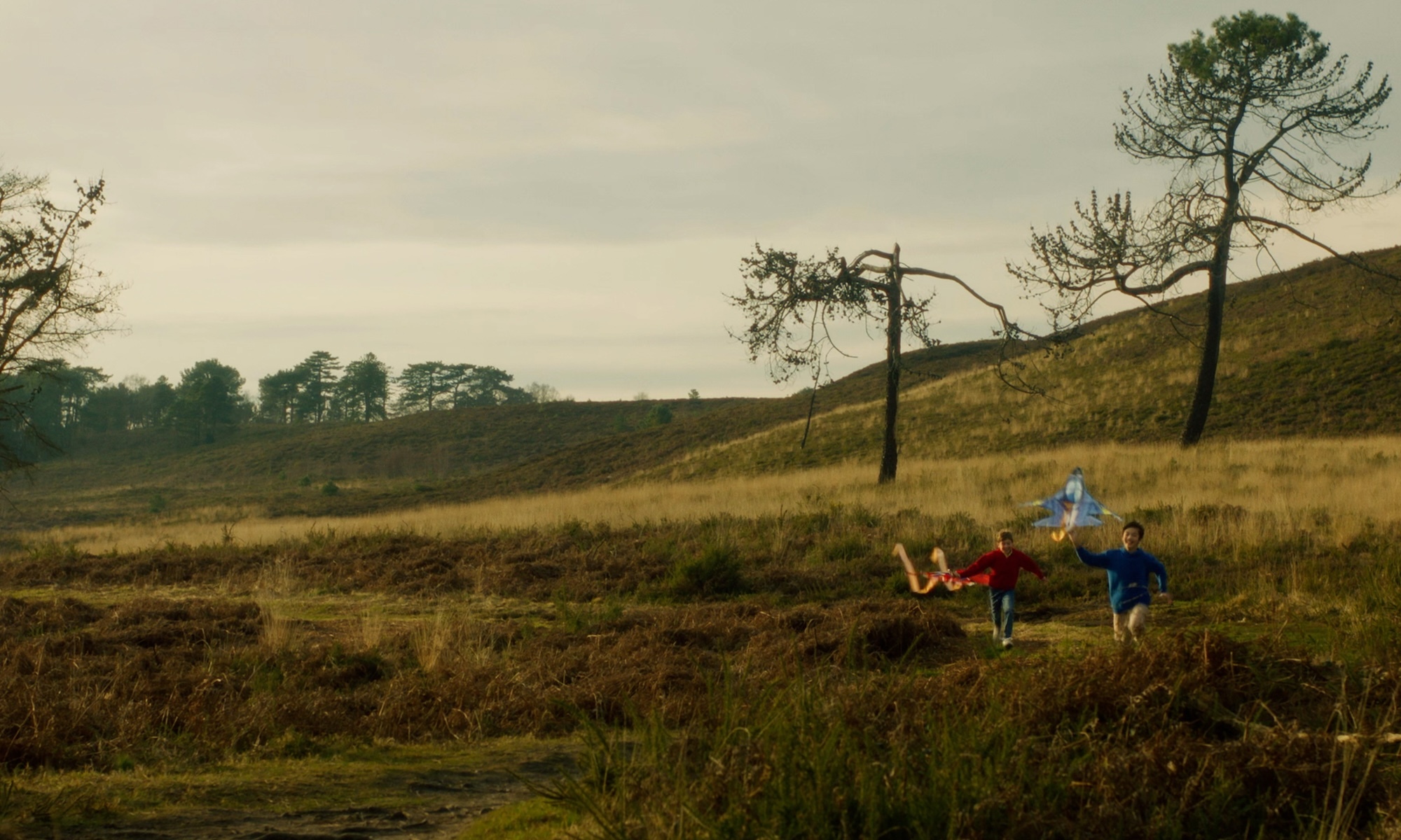 Two young people running with kites across moorland with scattered pine trees under overcast sky, rolling hills in background.