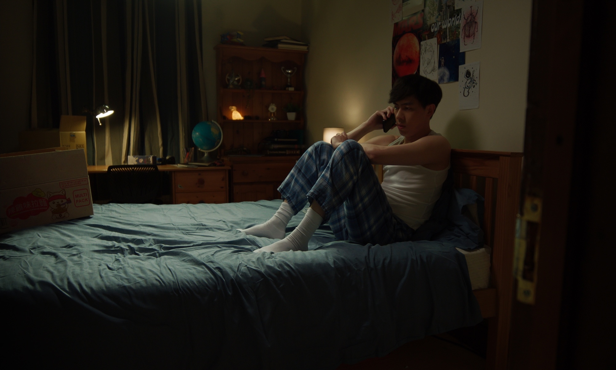Person in white vest and blue jeans sits on bed edge in dimly lit bedroom, holding mobile phone near ear.