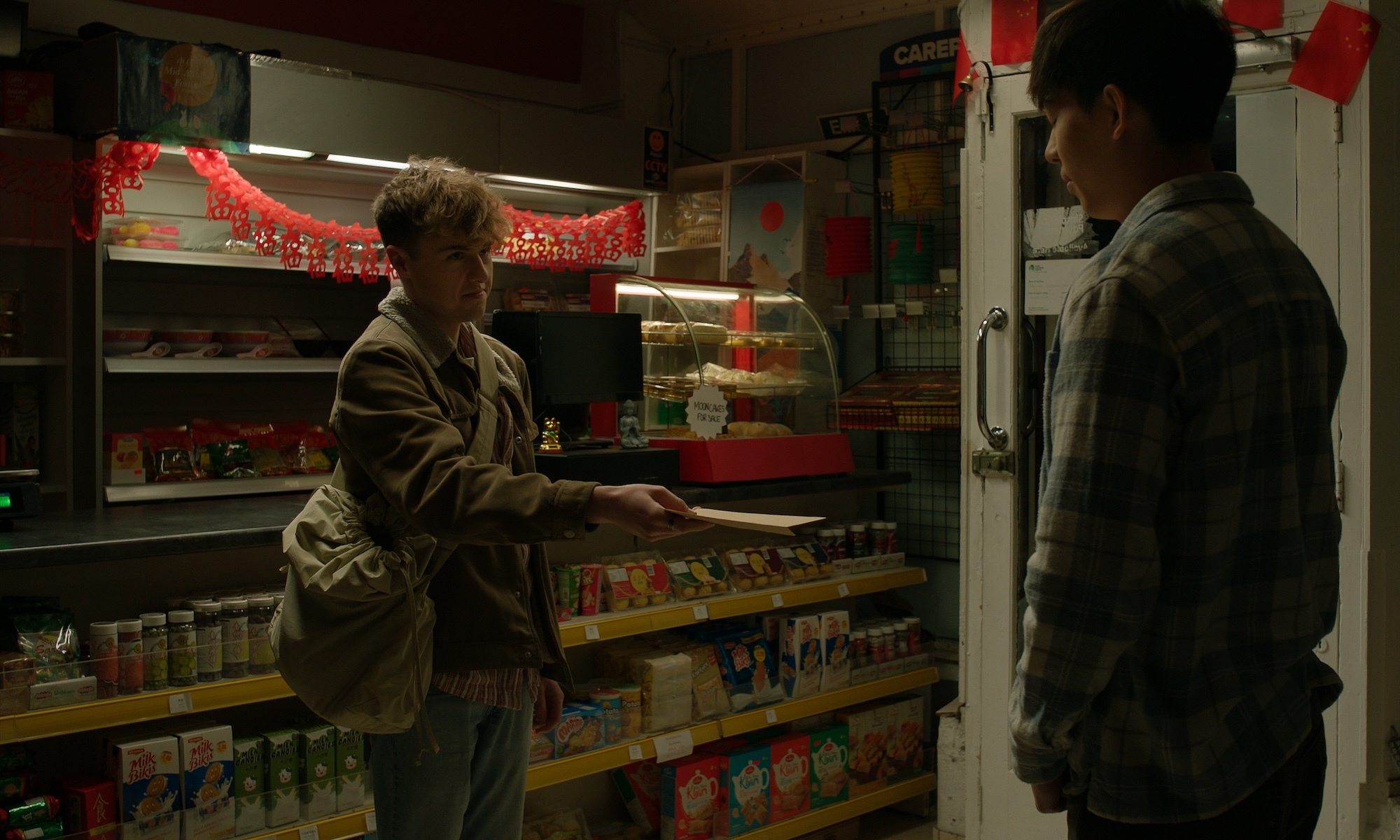 Two people in conversation inside a Chinese supermarket with shelves of products, red decorations hanging from ceiling, warm lighting.