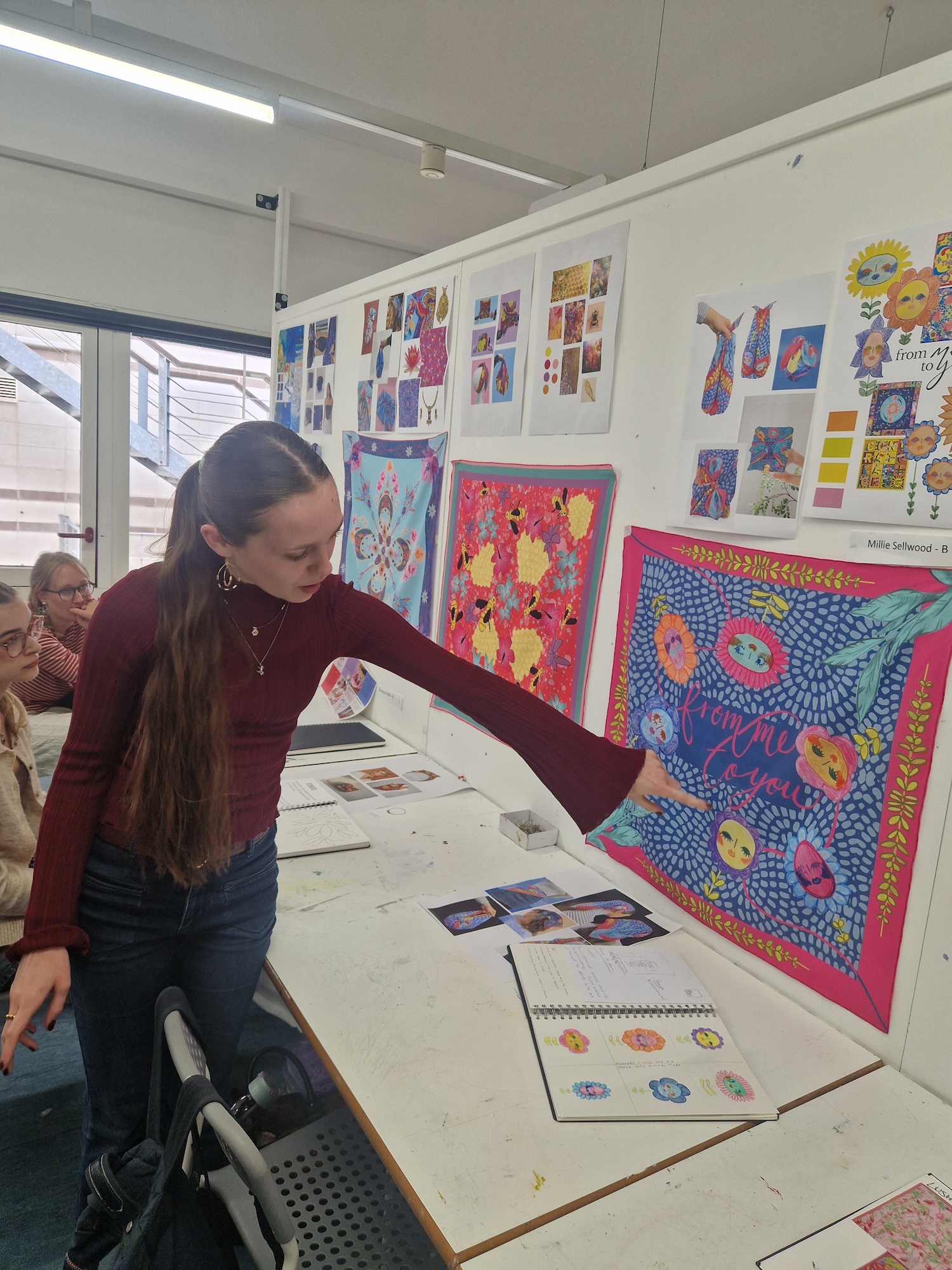 BA (Hons) Textiles Design student M Sellwood pointing at a bright and colourful textile design displayed on a studio wall.
