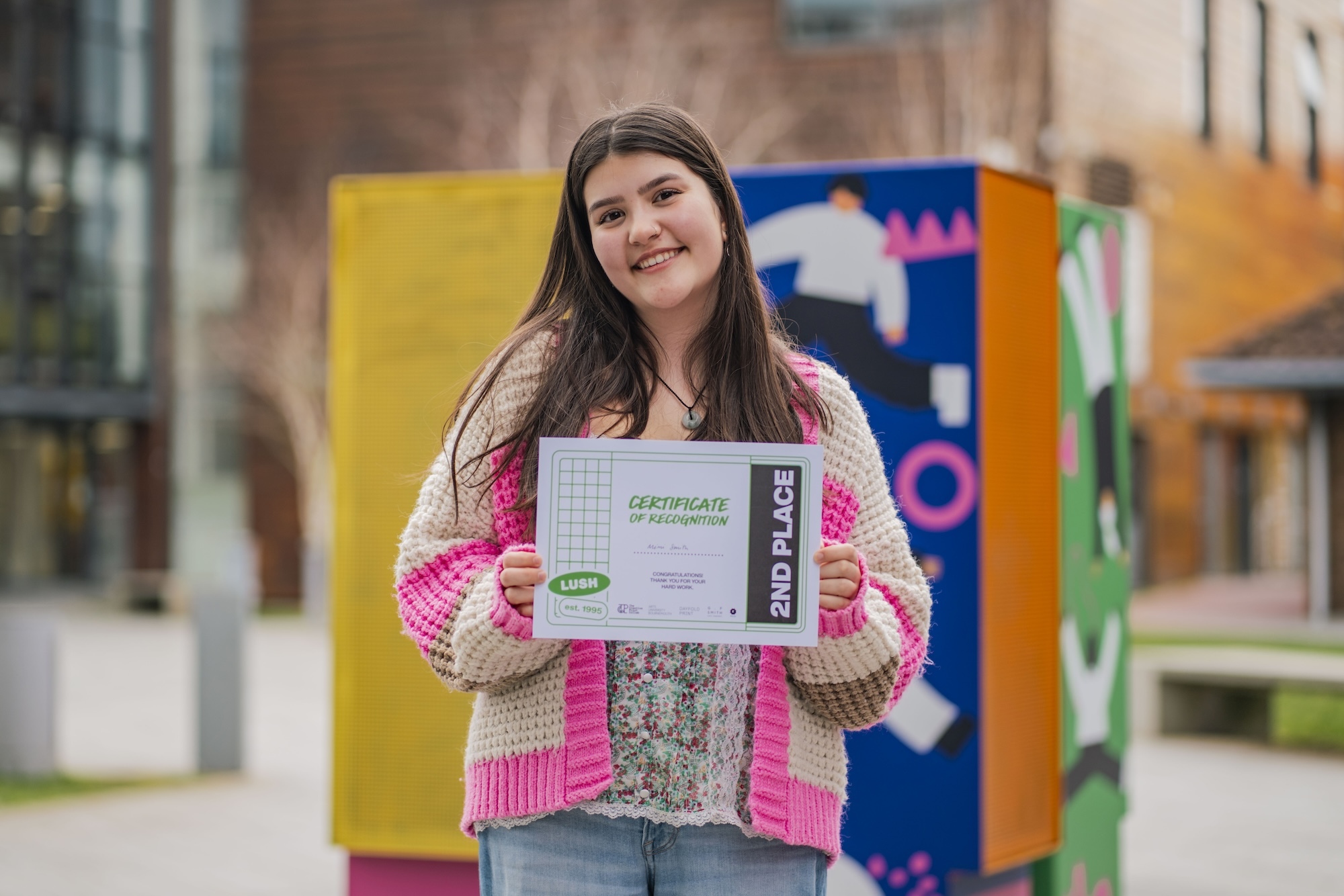 BA (Hons) Graphic Design student M Smith, holding a certificate for second place, posing for a photo in the courtyard outside University House, Arts University Bournemouth.