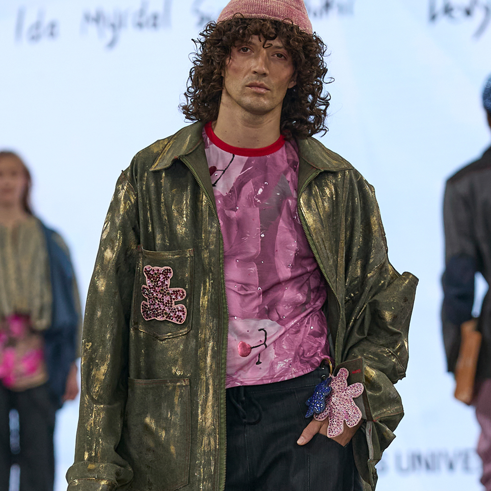 Male model on runway wearing burgundy beanie, metallic olive green jacket with patches, pink graphic t-shirt, and dark trousers.