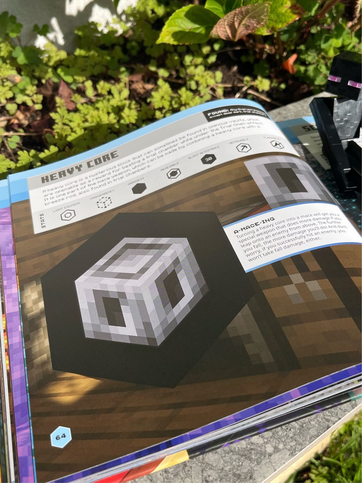 Open Minecraft guidebook showing pixelated cube structure on brown terrain with green foliage visible in background.