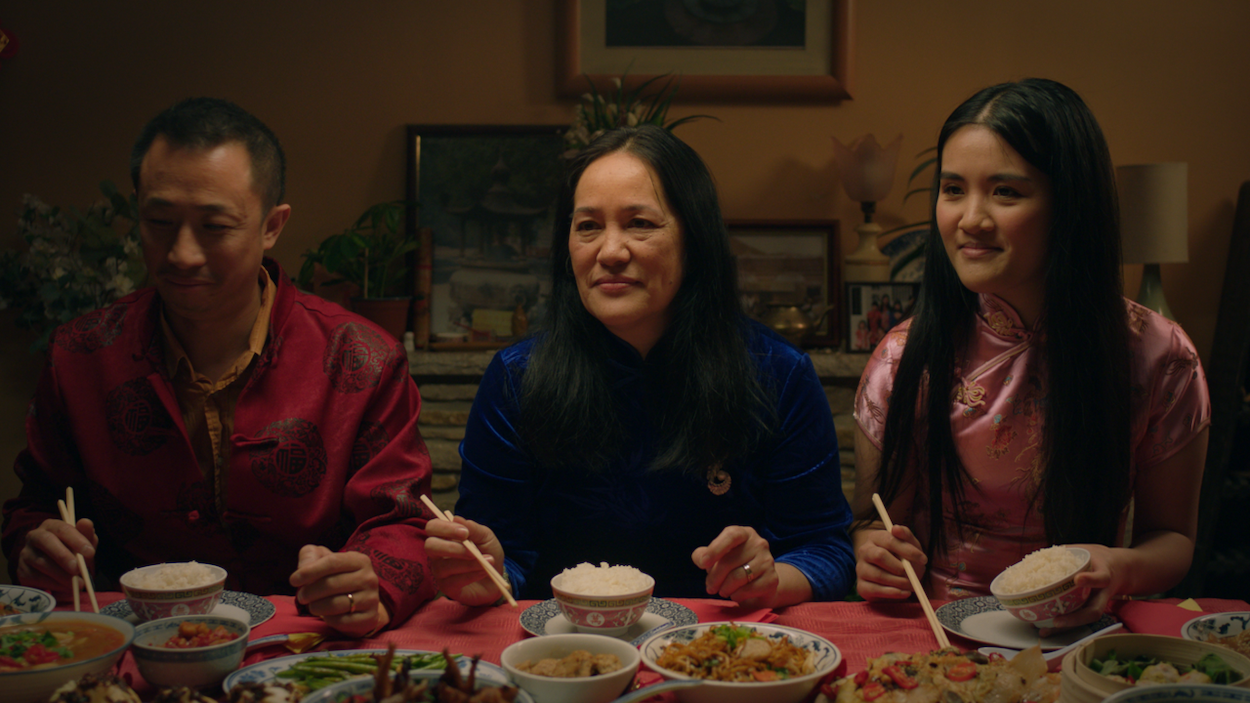 Three people sitting at dinner table with bowls and dishes, man in red shirt, older woman in blue, younger woman in pink top.