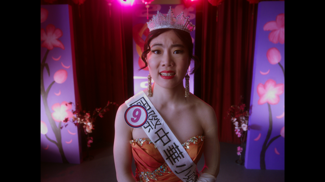 Woman wearing silver crown and white sash with Asian text, orange strapless dress, red curtains and purple lighting in background.