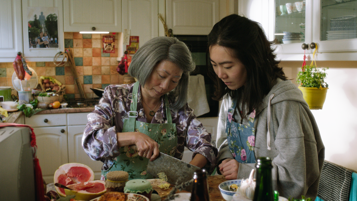 Two women preparing food together in kitchen with white cabinets, multicoloured tile backsplash, fresh vegetables and herbs on worktop.