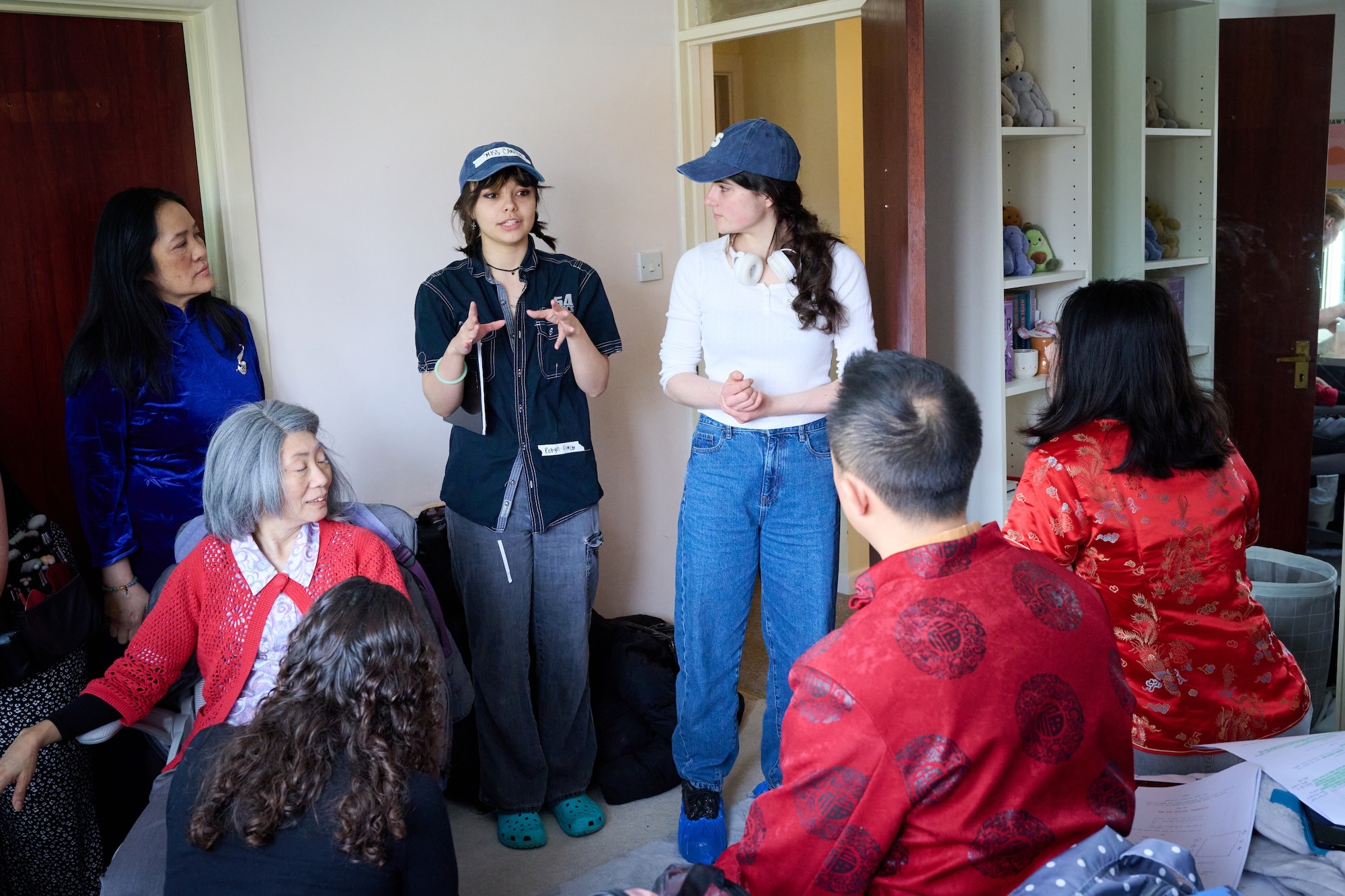 Two women in casual clothes and caps stand addressing seated elderly people in bright red clothing in a residential room with shelving.