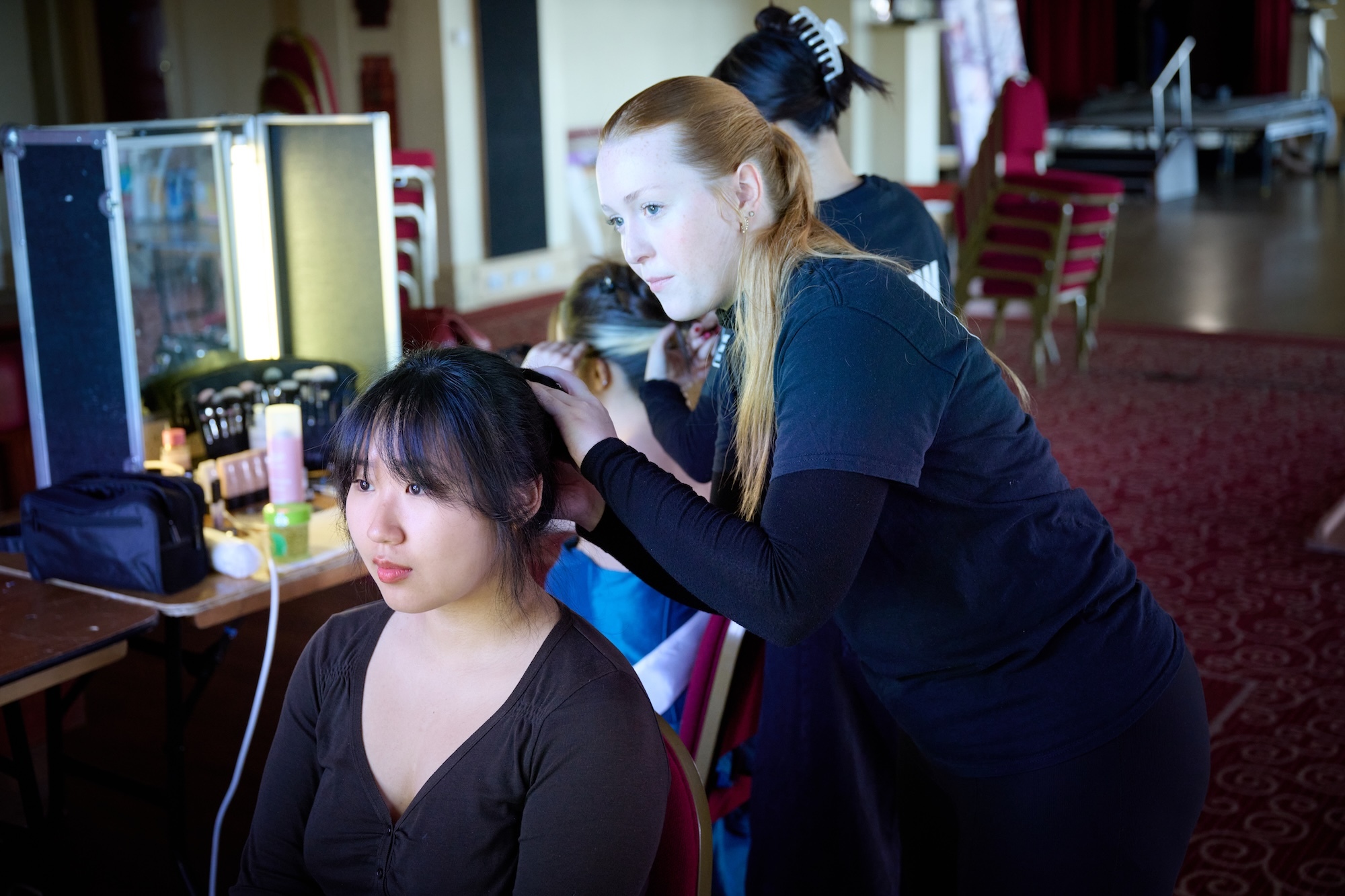 Hairdresser in black styling client's dark hair in salon with mirror, computer, and red carpet visible in background.