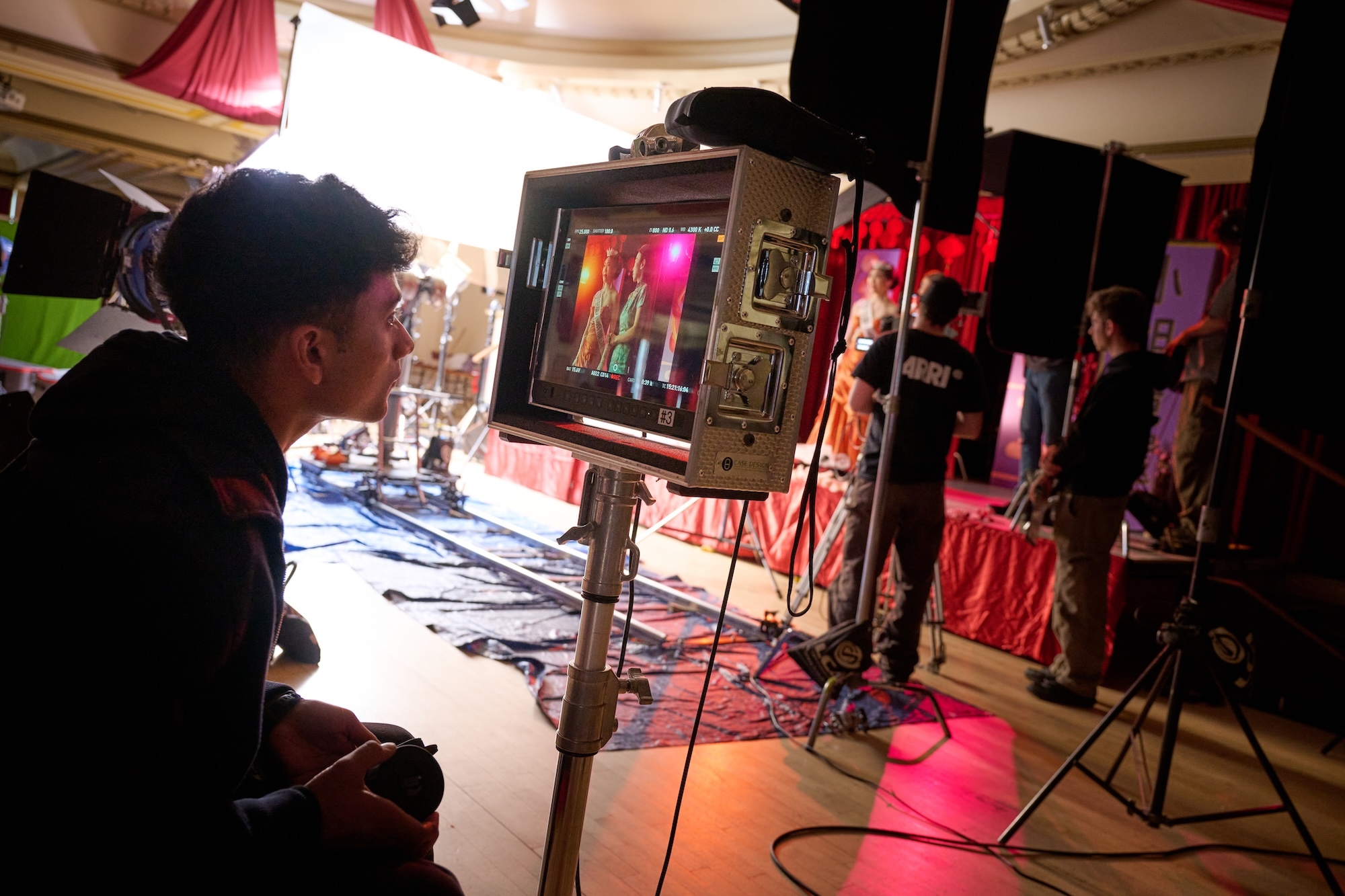 Film crew member operates camera equipment on set with red lighting and draped fabric backdrop, other crew visible in background.