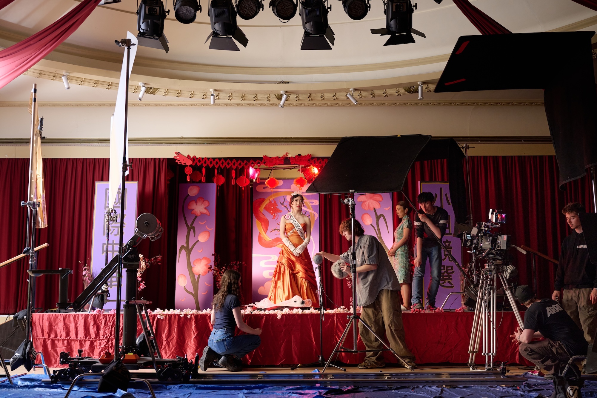 Stage set with red curtains, golden performer in centre, crew with cameras and lighting equipment, ornate ceiling with spotlights.