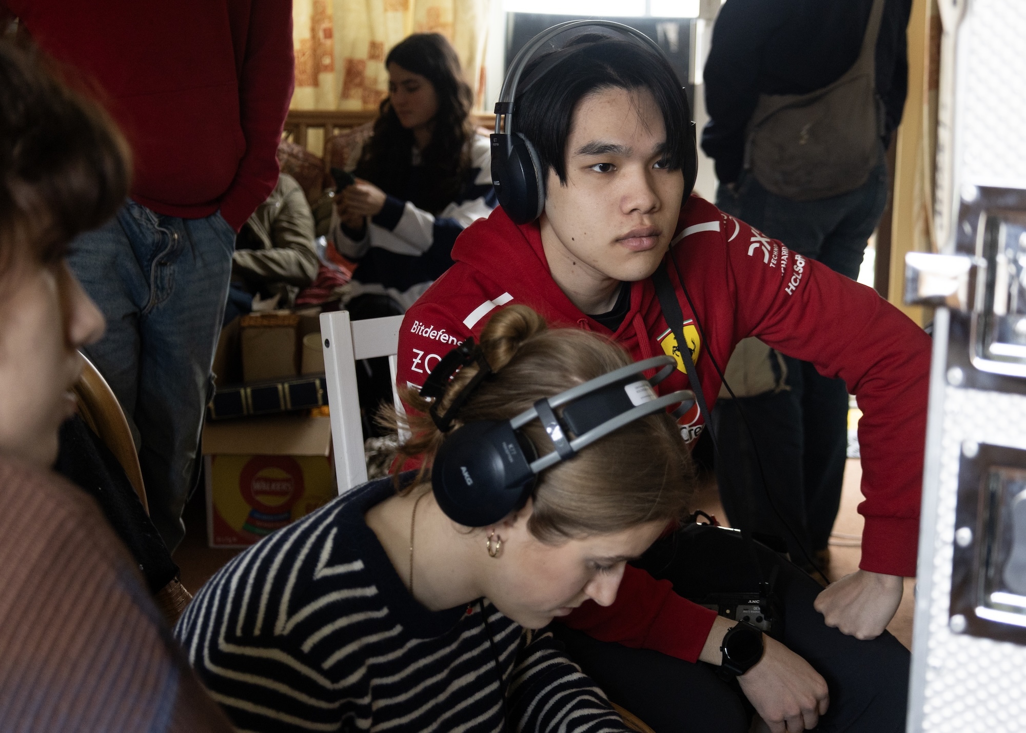 Person in red jumper using black hair clippers on someone in striped top seated in chair. Indoor setting with people in background.