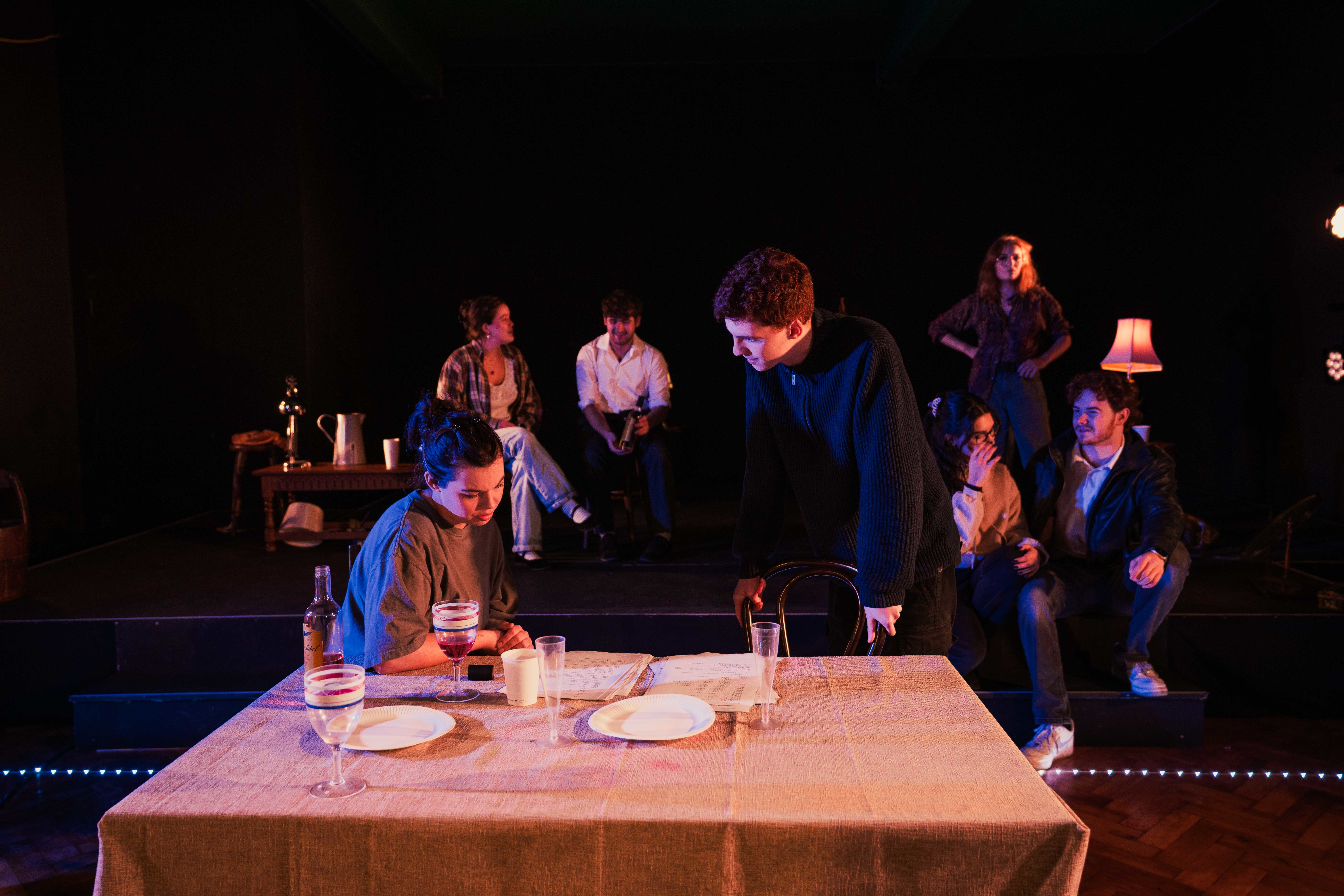 Two Acting students in conversation around a small square table. Other actors are visible talking among themselves in the dark background.