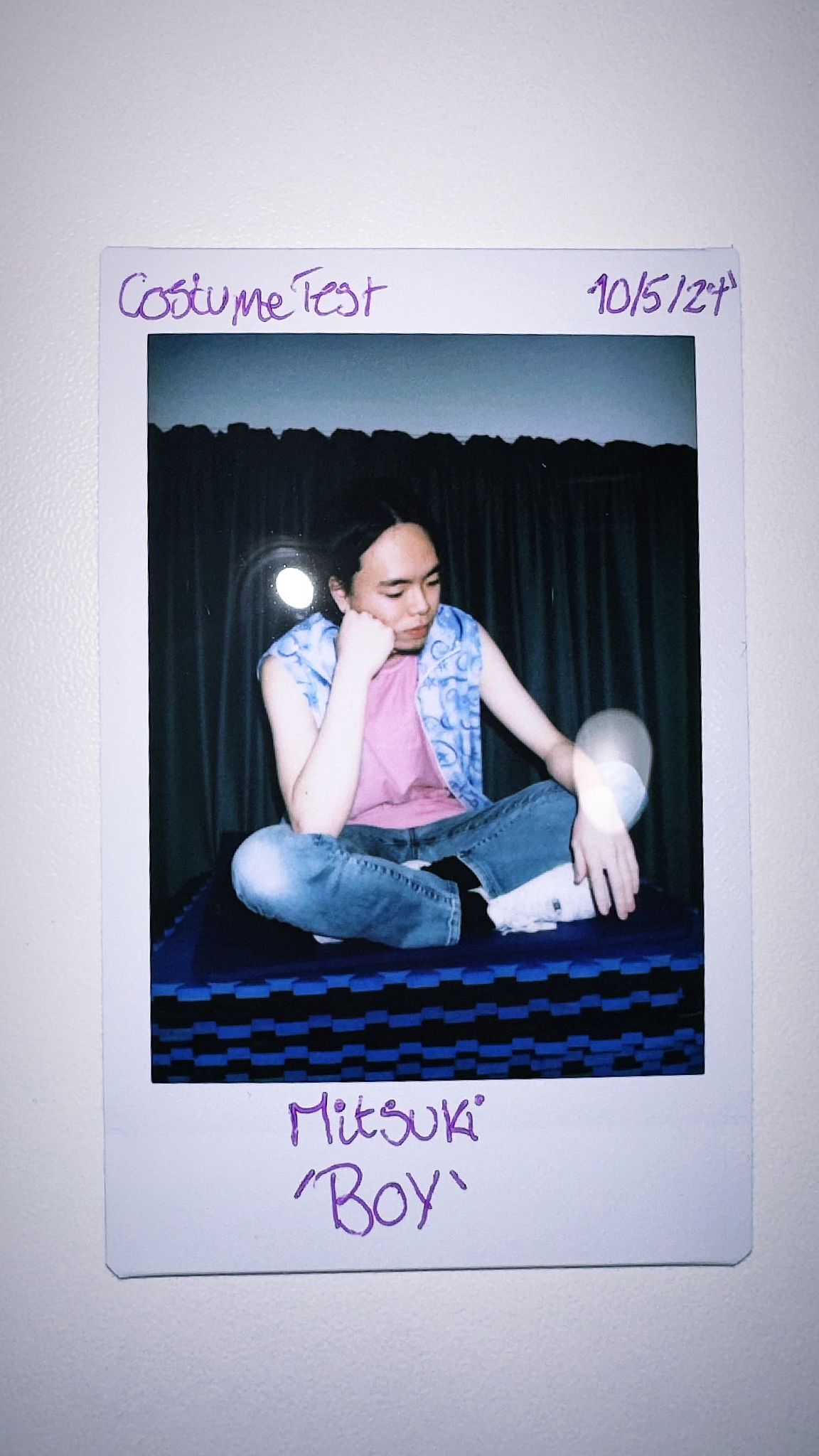 Polaroid shot of Mitsuki Kawase sat in front of a black curtain and lights, purple handwriting reads 'costume test, m