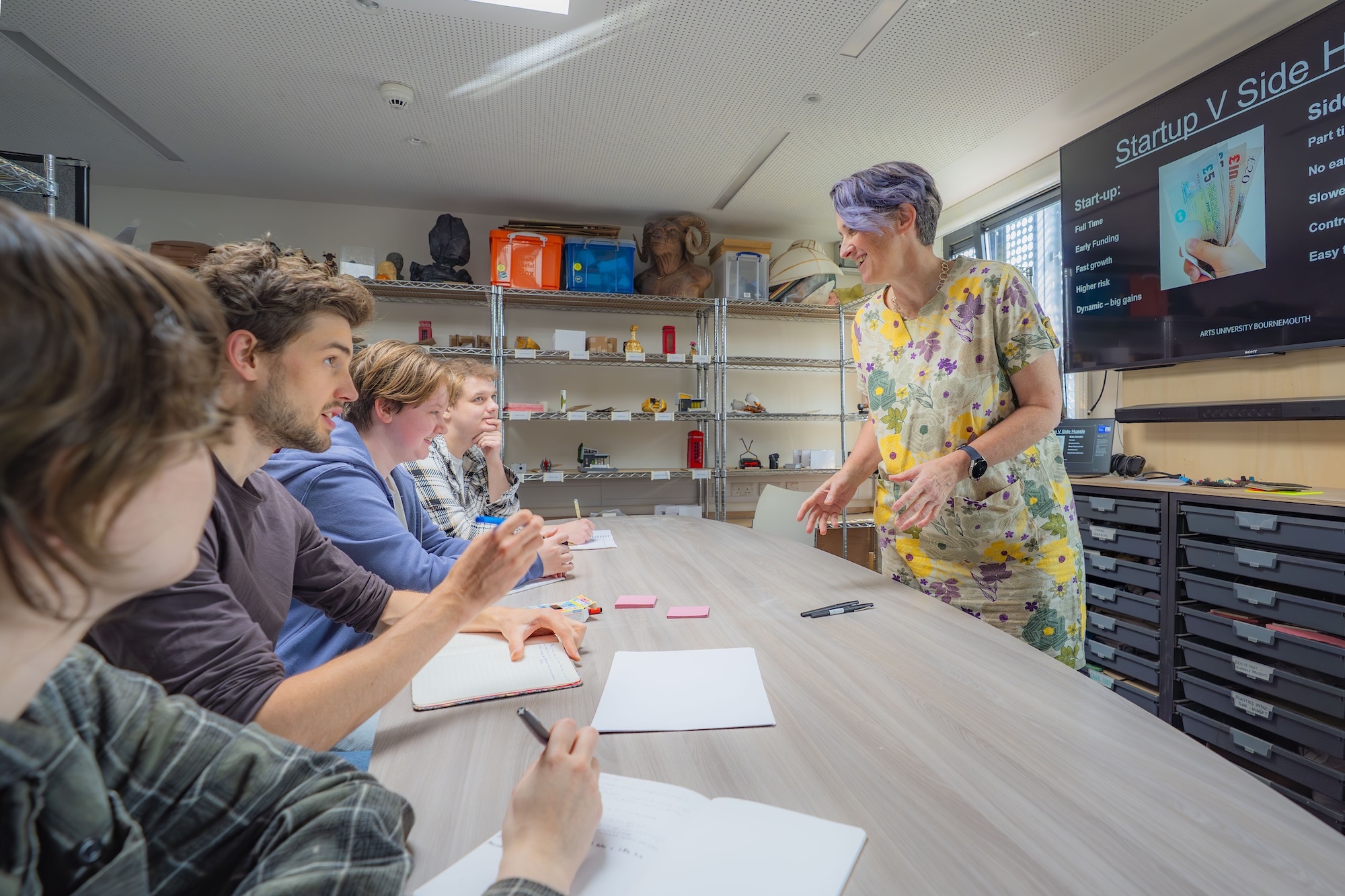Teacher in floral dress standing beside table with four seated students working on papers in classroom with shelves and whiteboard.