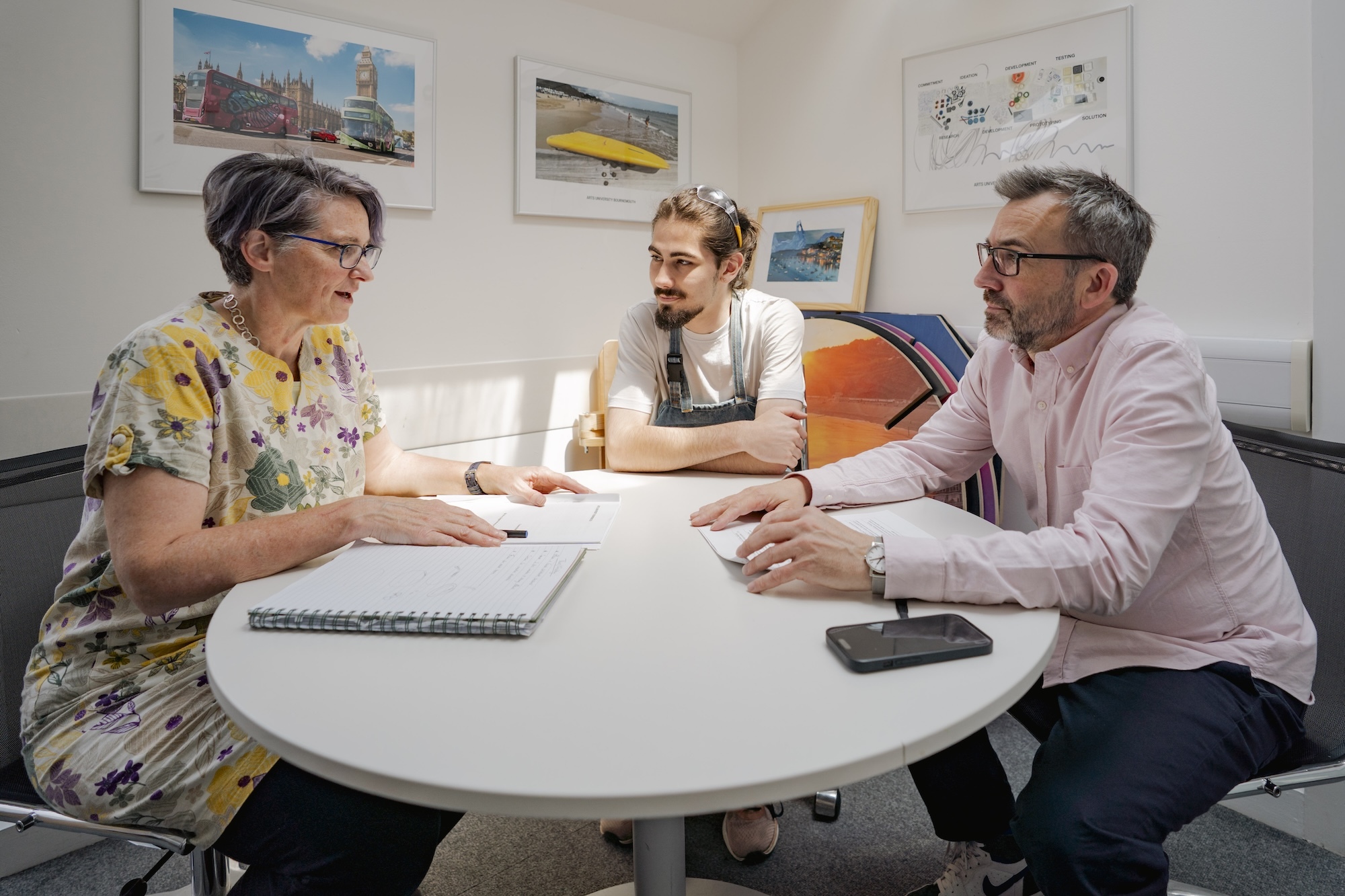 Three people sitting at white round table in meeting room, woman in floral top with notebook, man and woman reviewing documents together.