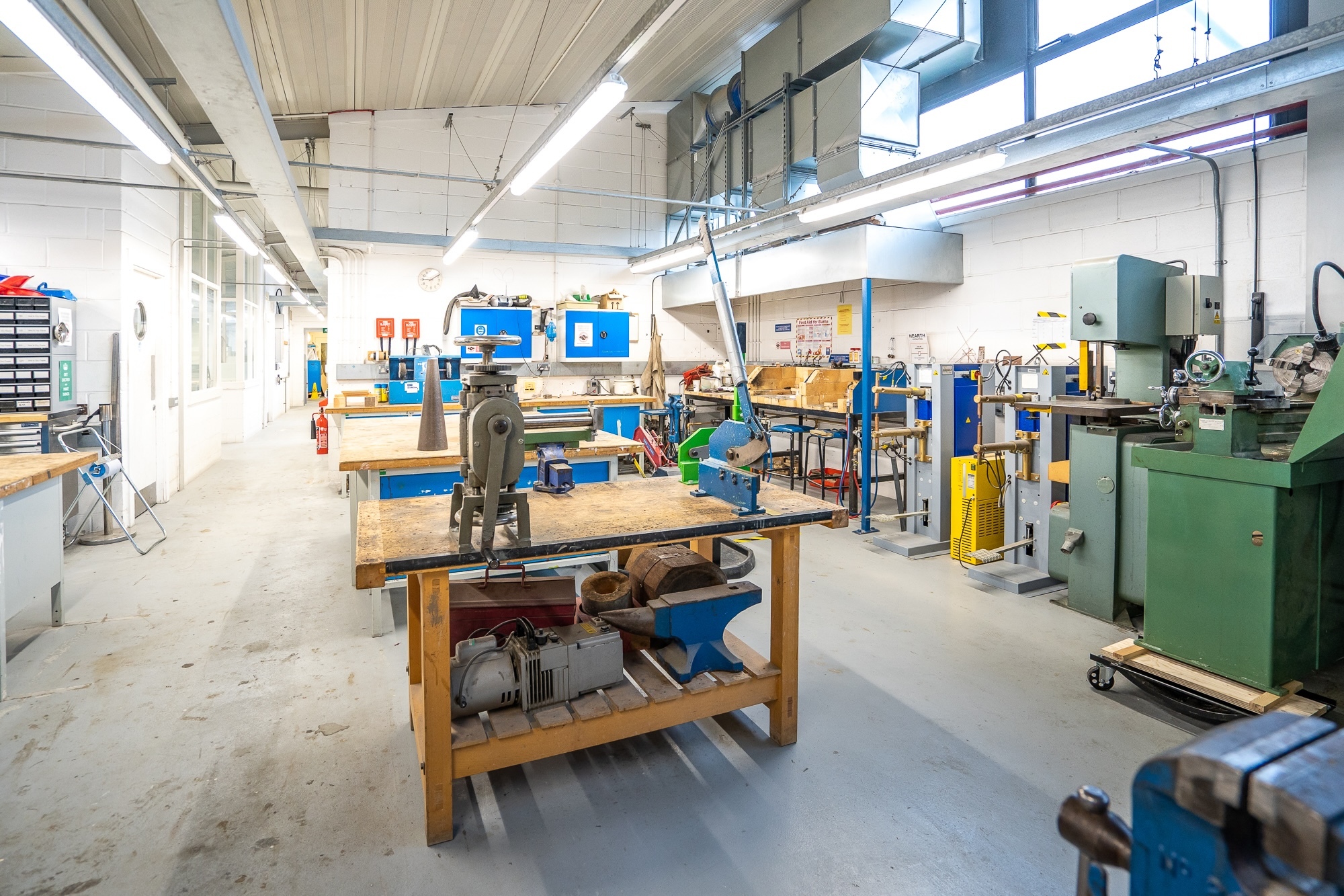 Industrial workshop with wooden workbenches, blue and green machinery, white walls, exposed ceiling with fluorescent lighting.