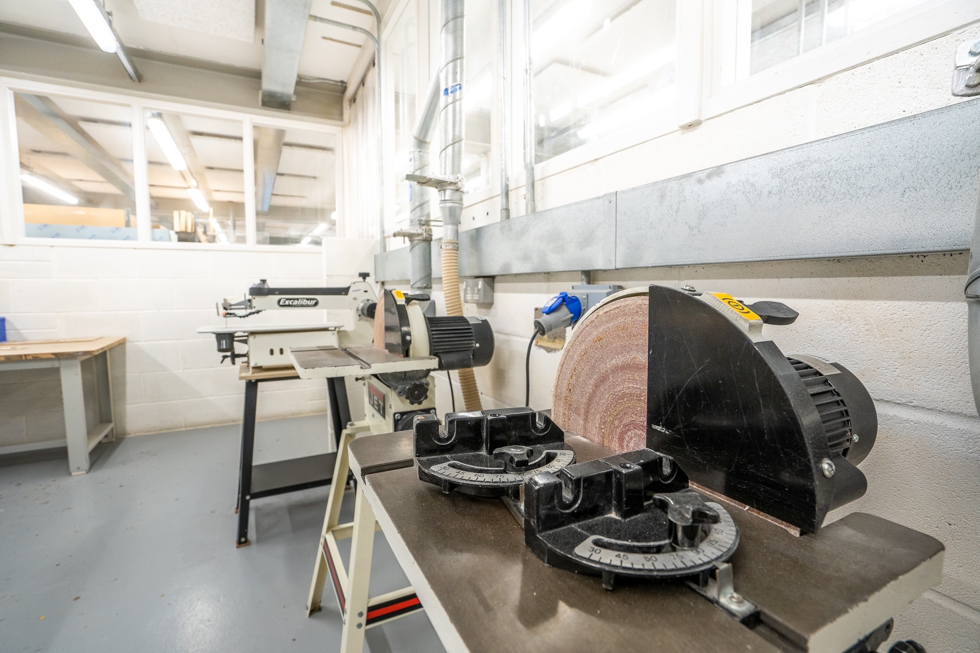 Industrial workshop with white walls, concrete floor, metal workbenches, and various machinery including a large circular saw and cutting equipment.