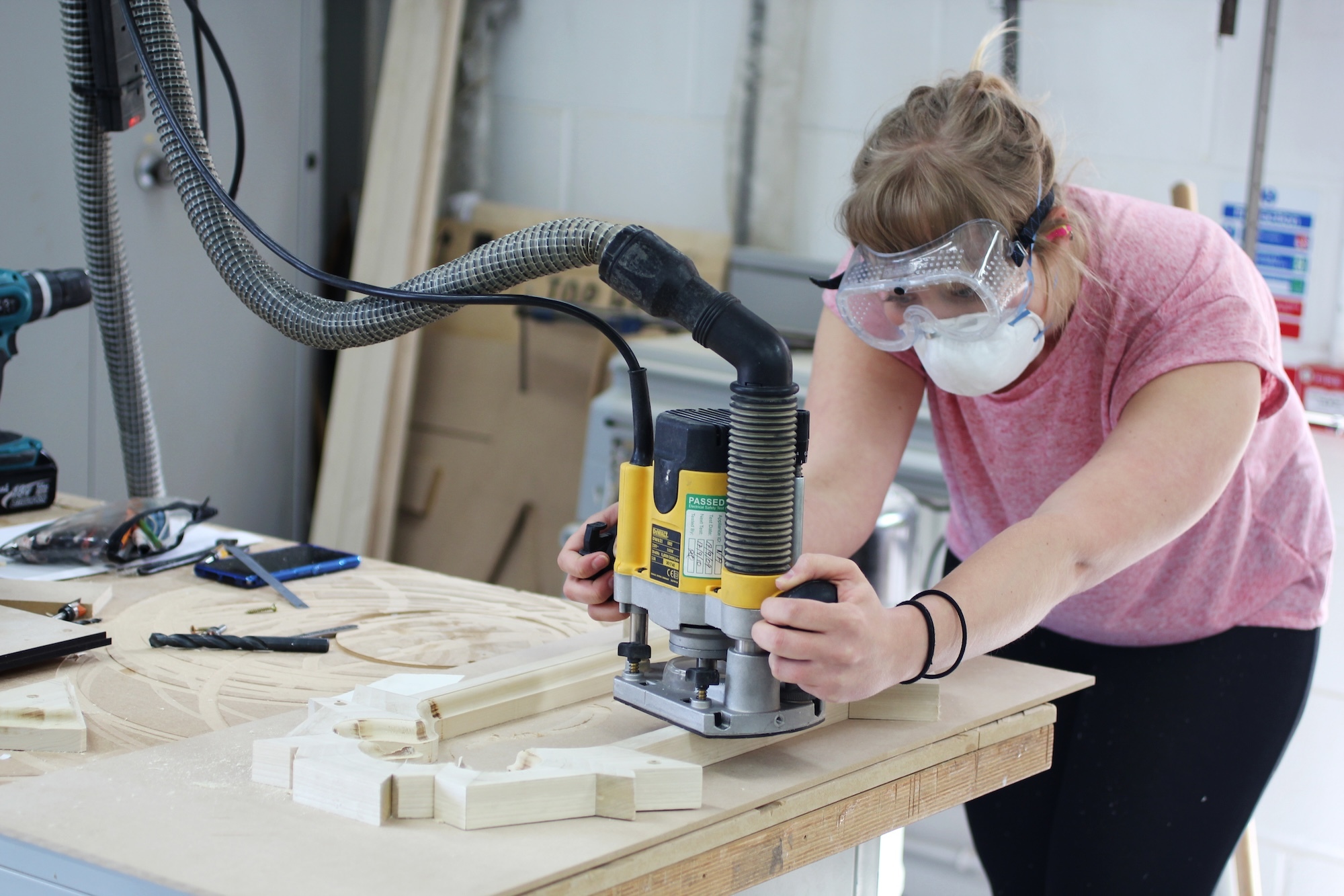 Woman in pink shirt and safety mask using yellow router with dust extraction system on wooden workbench in workshop.