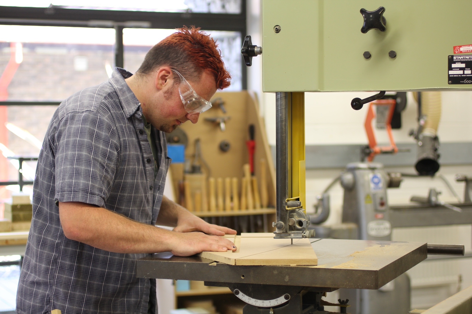 Man in grey checked shirt operating bandsaw on wooden board in workshop with tools and equipment visible in background.