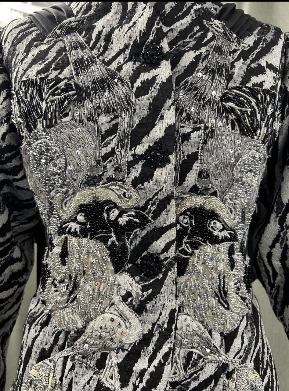 A close-up view of an intricately embroidered garment featuring a black and white zebra pattern. The design showcases detailed images of birds and floral motifs, embellished with beads and sequins for added texture.