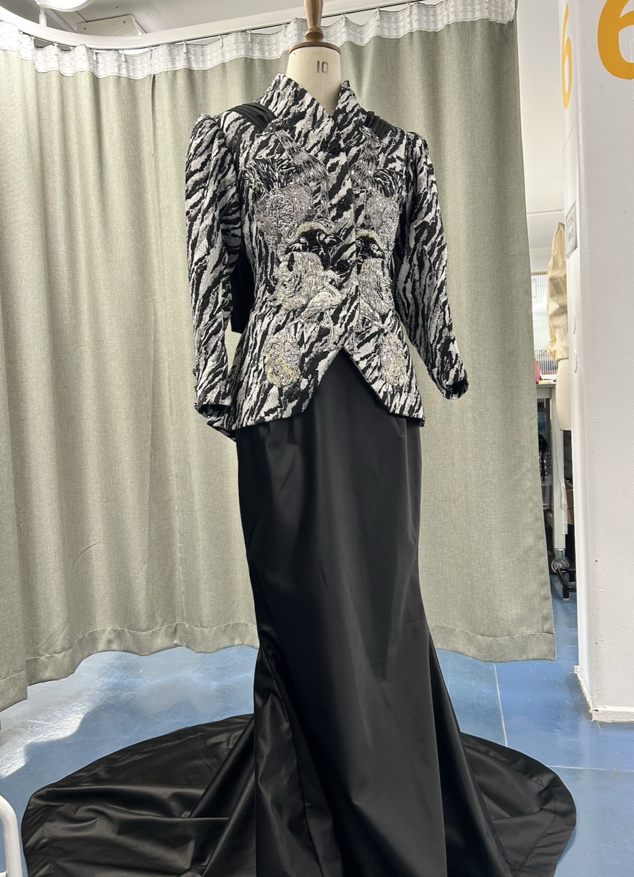 A mannequin displays a black and white jacket with a textured zebra print pattern paired with a long black skirt that has a train. The background features draped fabric in a muted colour and a blue floor.