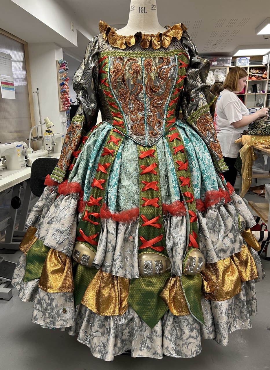A vibrant historical dress displayed on a mannequin in a sewing workshop. The dress features intricate patterns and textures, with colours including turquoise, green, red, and gold, complemented by ruffles and decorative bells.