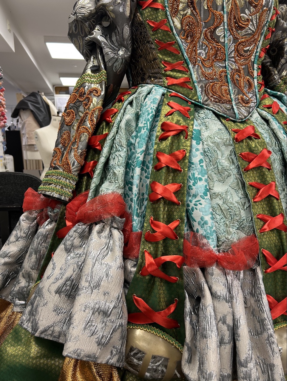 A detailed close-up of a multi-layered costume featuring vibrant fabrics in green, blue, and red. The dress has intricate patterns, with red ribbons and ruffled sleeves complementing the ornate design. The background indicates a workshop or garment display.