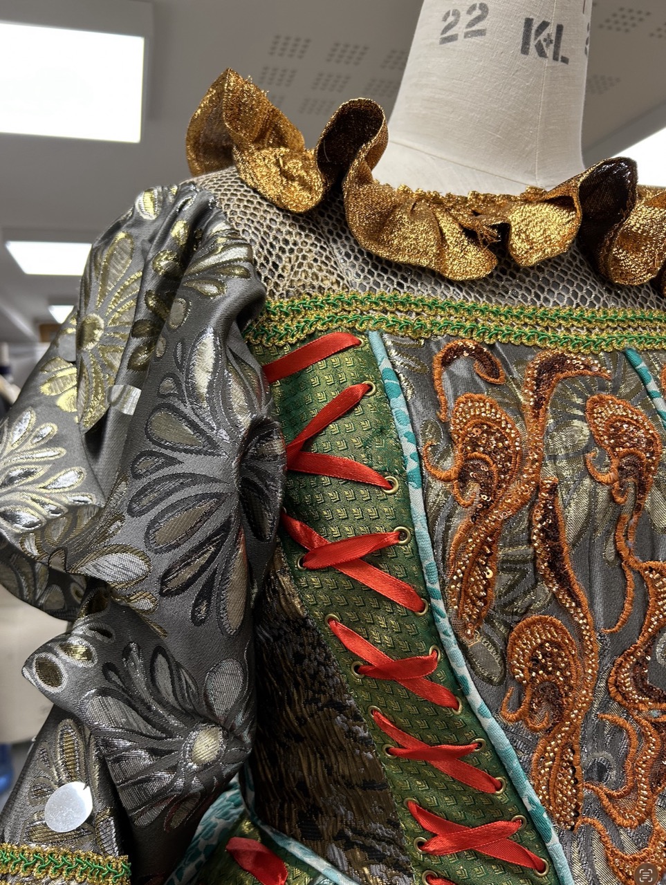 A close-up of a dress with intricate detailing, featuring a metallic grey fabric adorned with golden ruffles and red embellishments. The bodice displays ornate patterns with beads and embroidery, showcasing vibrant colours and textures.