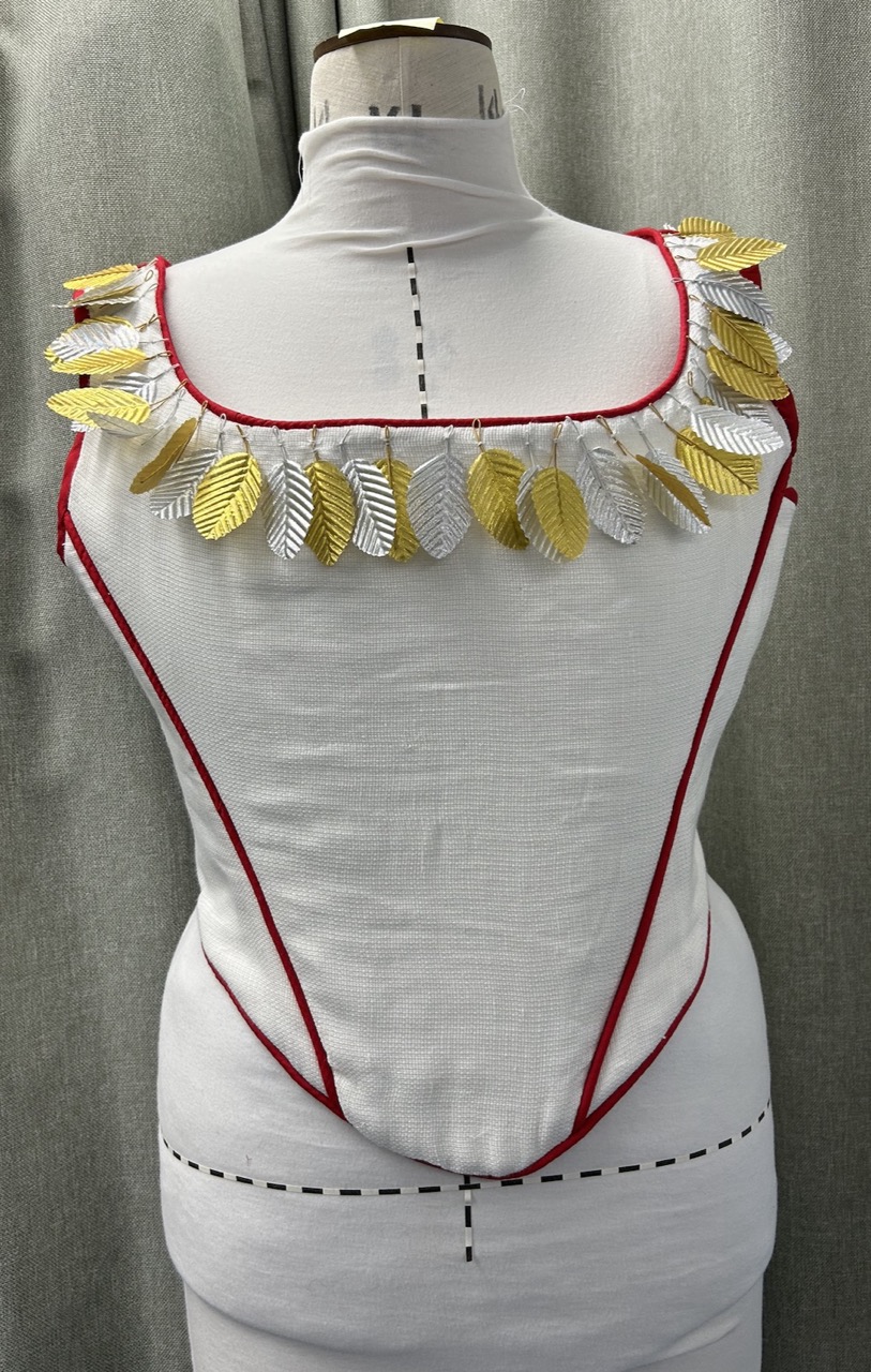 A white fabric garment displayed on a dress form, featuring red trim and a decorative neckline adorned with metallic gold and silver leaf-shaped appliqués. The background is a soft grey, providing contrast to the garment.