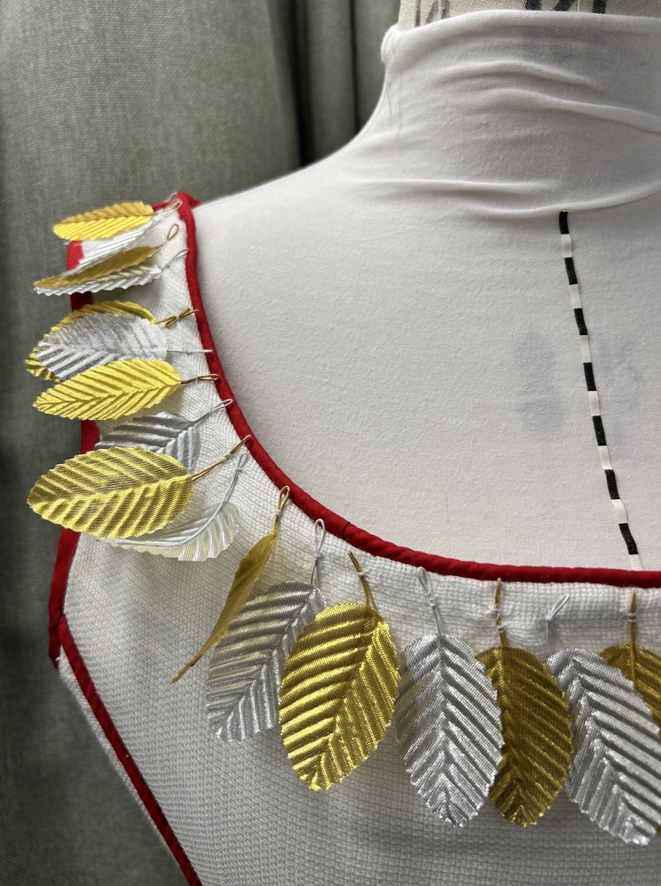 Close-up of a fashion garment featuring a collar adorned with alternating silver and gold leaf-shaped embellishments. The collar is edged with red trim, and the garment is displayed on a white dress form against a textured grey background.