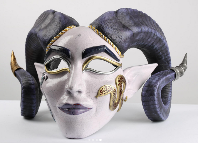 A decorative white mask with gold details and dark curled horns, displayed against a plain light background.