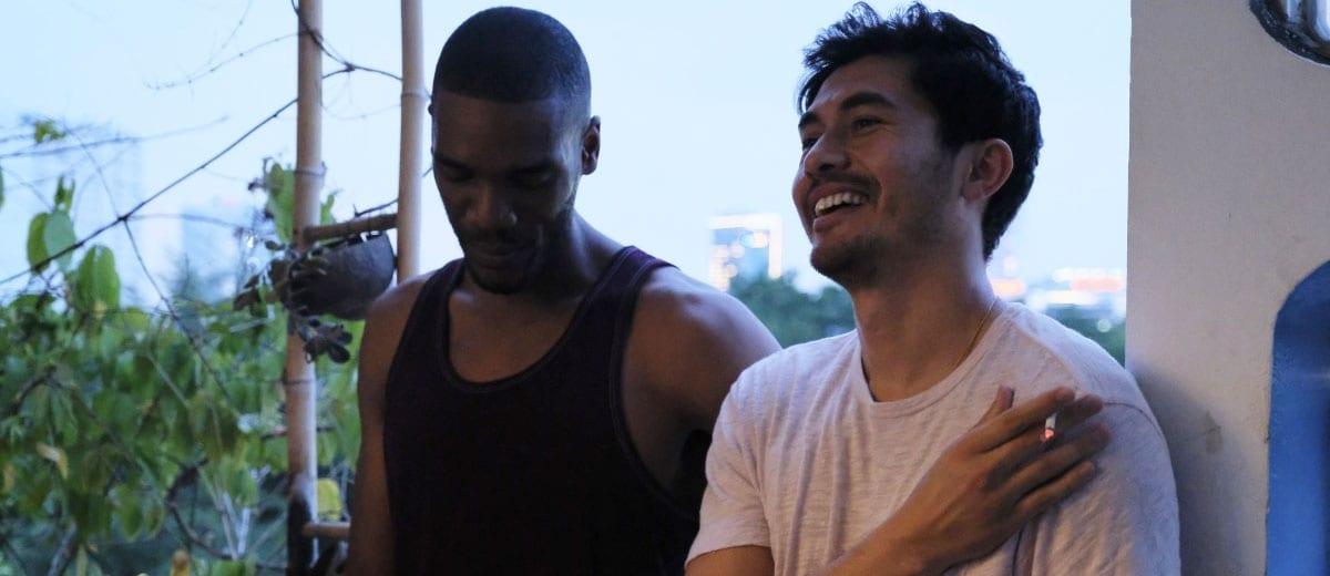 Two men standing together. One is leant up against a pillar smiling with a cigarette in his hand.