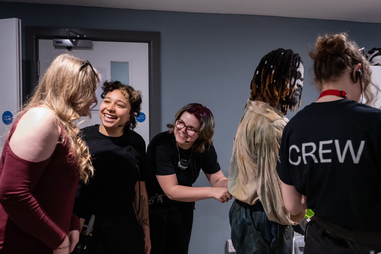 Four crew members stand in a backstage area, with one wearing a black shirt marked CREW and another in a beige fringed jacket.