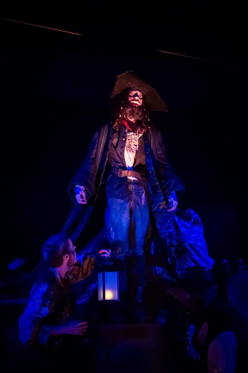 A life-sized pirate figure in a tricorn hat and long coat stands above people below, lit in blue and red light against a dark background.