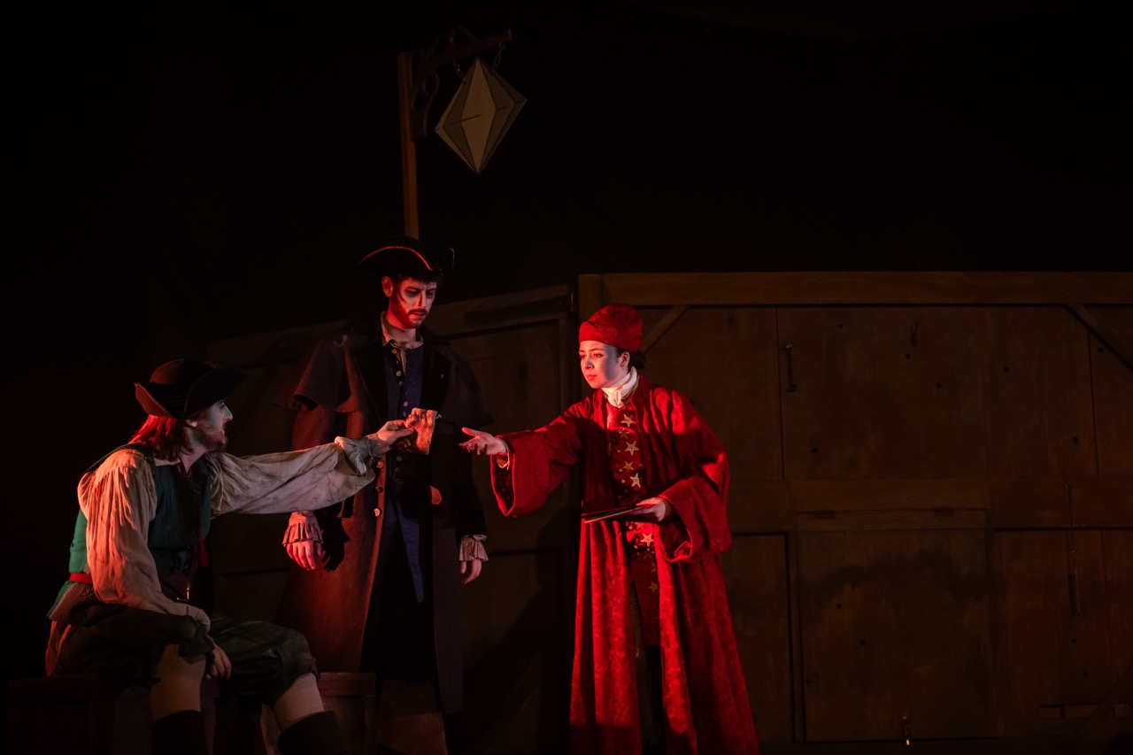 Three actors in period costumes perform on a dimly lit stage, with one seated at left and two standing centre and right against a wooden wall.
