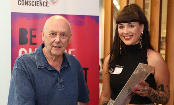 Two people standing and posing for a photo. Person on the right is holding an award.