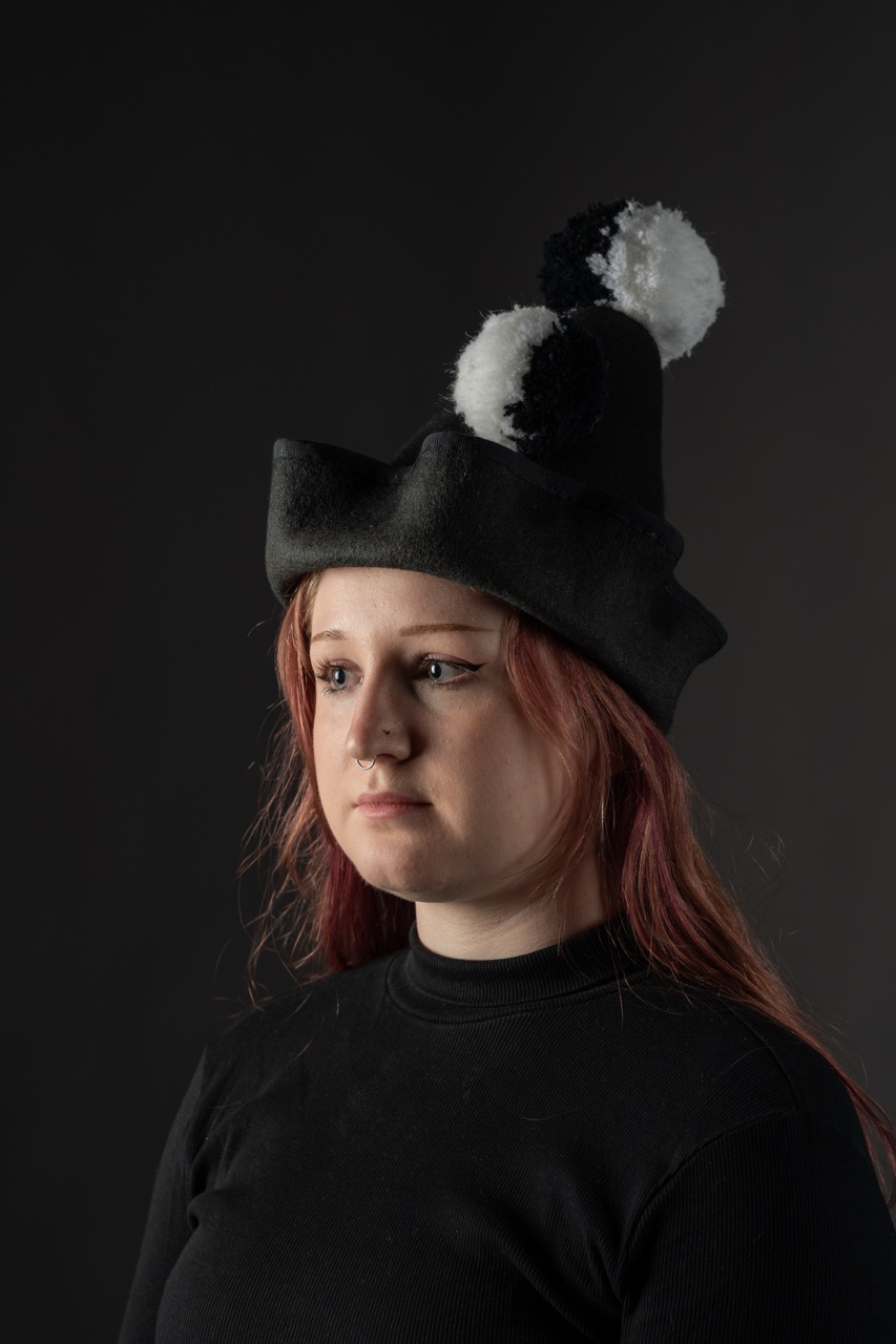 A woman with long red hair wears a black hat adorned with white and black pom-poms. She is dressed in a black top, and the background is dark, creating a contrast with her lighter features.