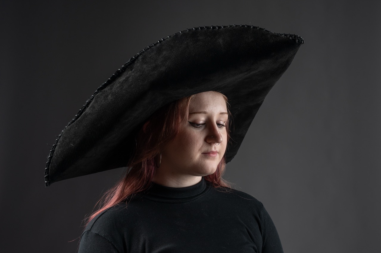 A young woman with shoulder-length, reddish hair wears a large, black hat with a wide brim. She is dressed in a black top, and the background is a dark grey, creating contrast with the hat.