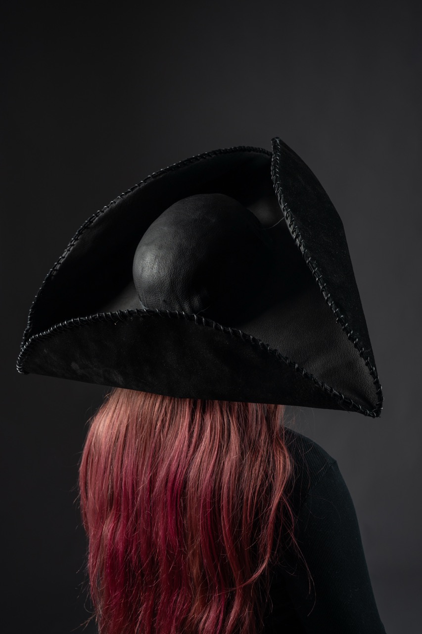 A person with long hair featuring pink highlights is seen from behind, wearing an oversized black hat with an angular design and a rounded top. The background is a dark grey, enhancing the hat's distinctive shape.
