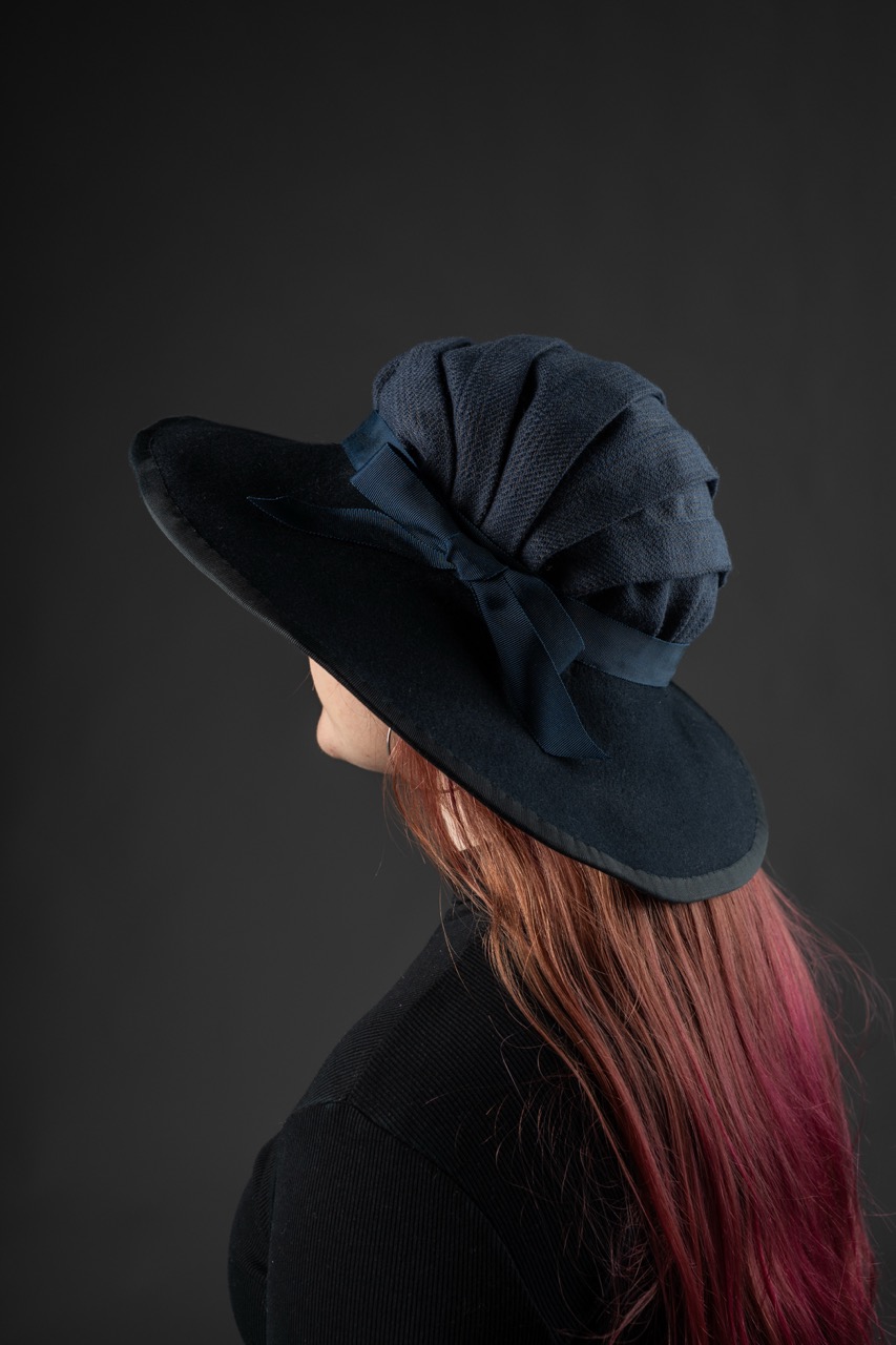 A woman with long hair, tinted red at the ends, is wearing a large-brimmed navy hat with a pleated design and a ribbon. The photograph has a black background, highlighting the hat's features.