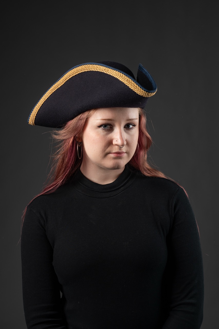 A young woman with long red hair wearing a black turtleneck sweater poses against a dark background. She is adorned with a large, navy blue tricorn hat accented with gold trim.