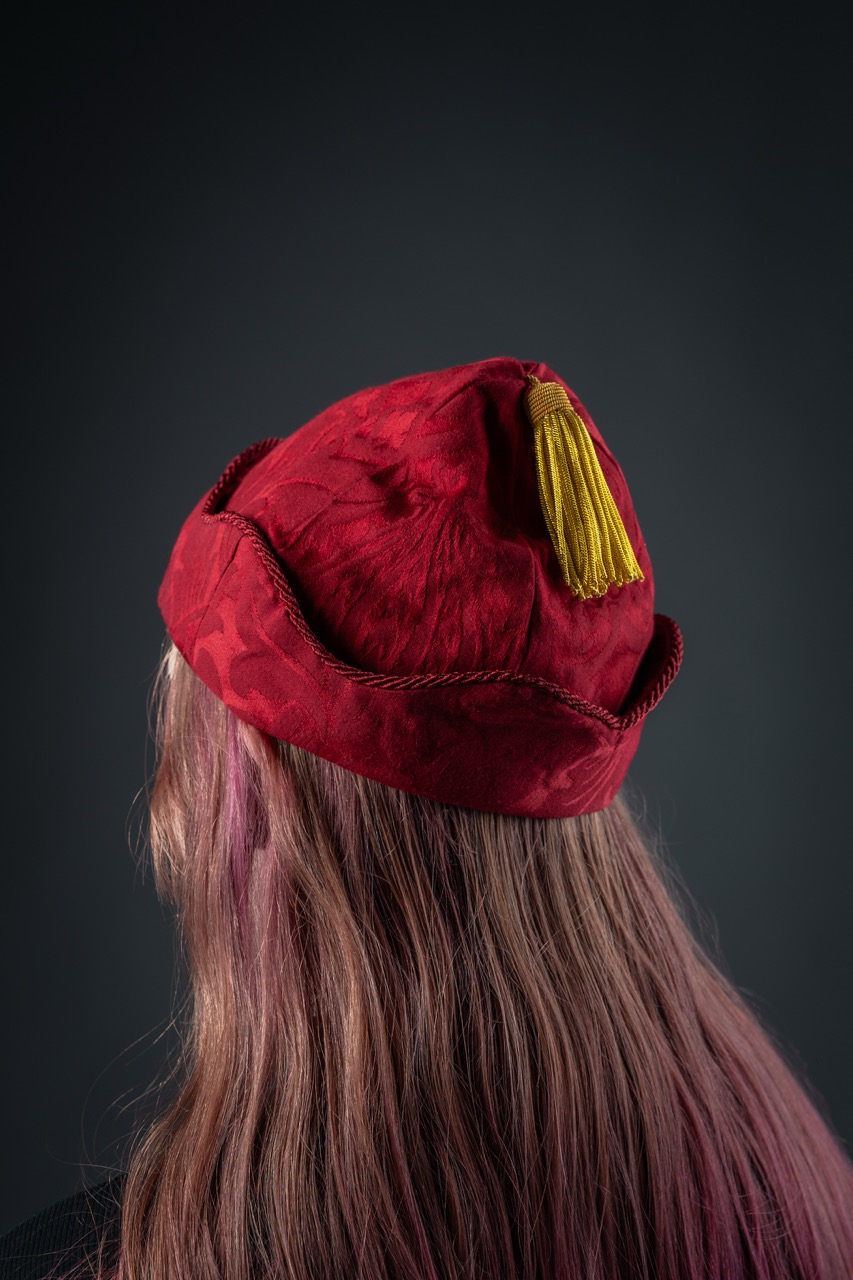 A close-up view of a person’s head from behind, featuring long hair with pink highlights. They are wearing a red academic cap, adorned with a yellow tassel, against a dark grey background.